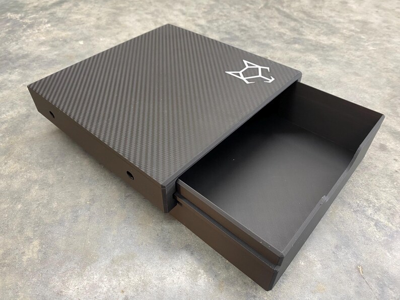 May include: A black drawer with a carbon fiber top and a white wolf logo. The drawer is open and the inside is black.