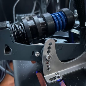 Puede incluir: Primer plano de un conjunto de pedales de simulador de carreras negro y plateado con un resorte azul y un cable negro.