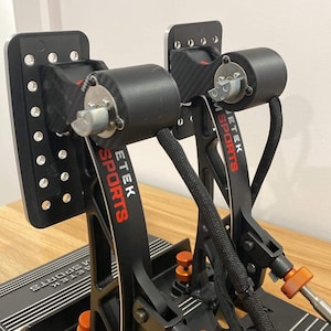 Puede incluir: Un par de pedales de carreras negras con botones de ajuste naranja. Los pedales están etiquetados con "ETEK SPORTS" en letras rojas. Los pedales están montados en una base negra con una franja plateada.