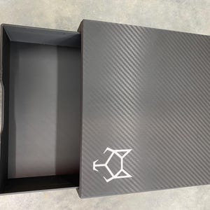 May include: A black box with a carbon fiber pattern and a white logo. The box has a drawer that is open, revealing an empty interior.