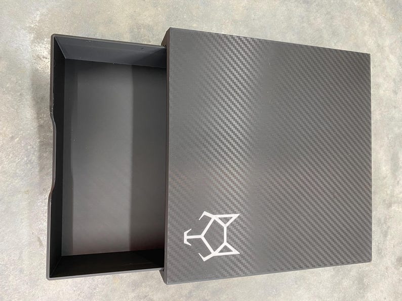 May include: A black box with a carbon fiber pattern and a white logo. The box has a drawer that is open, revealing an empty interior.