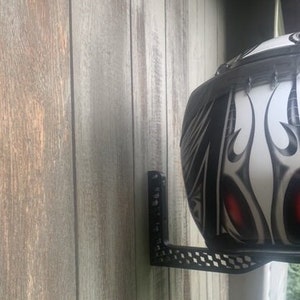 K&ouml;nnte beinhalten: Ein schwarzer Helmhalter aus Metall, der an einer Holzwand montiert ist. Der Halter hat ein Schachbrettmuster und eine vertikale St&uuml;tze. Ein wei&szlig;er Motorradhelm mit schwarzen und roten Tribal-Designs ist am Halter befestigt.