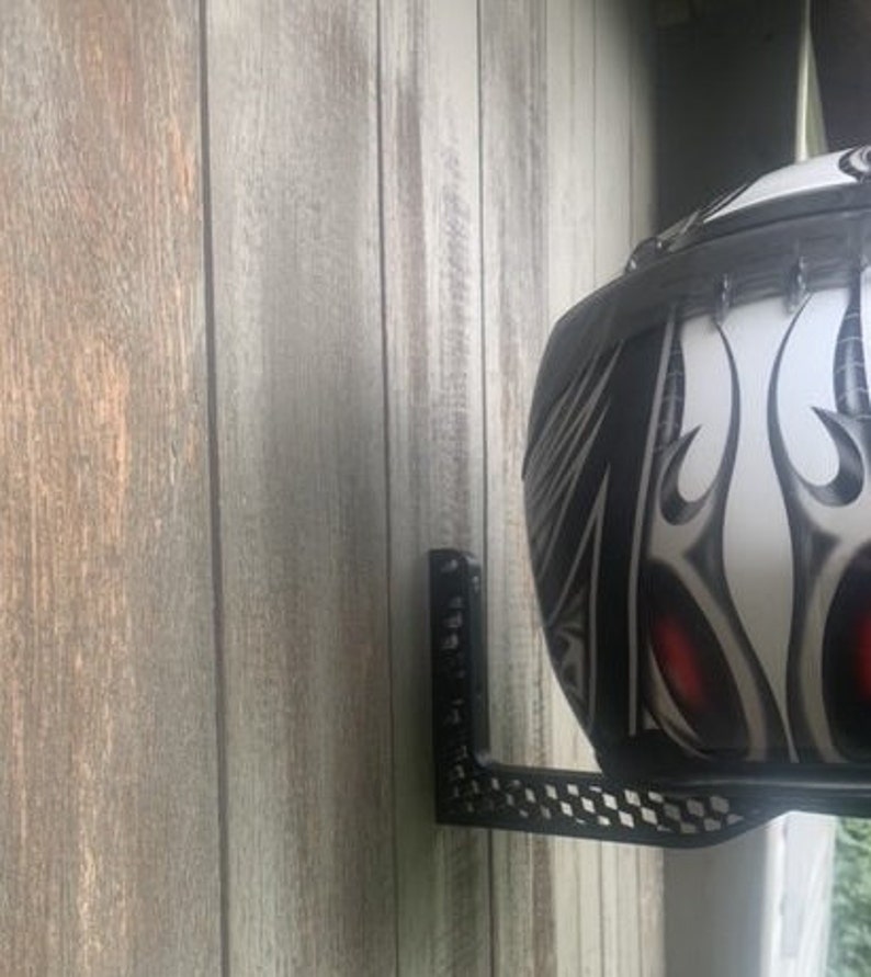 K&ouml;nnte beinhalten: Ein schwarzer Helmhalter aus Metall, der an einer Holzwand montiert ist. Der Halter hat ein Schachbrettmuster und eine vertikale St&uuml;tze. Ein wei&szlig;er Motorradhelm mit schwarzen und roten Tribal-Designs ist am Halter befestigt.