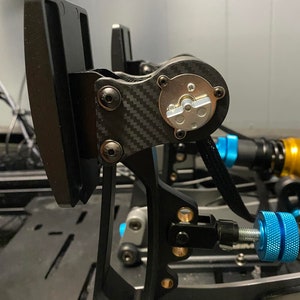 May include: Close-up of a black and silver racing pedal with a carbon fiber design. The pedal has a black metal arm with a gold-colored adjustment knob. The pedal is mounted on a black metal frame.