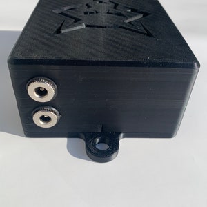 May include: A black 3D printed box with a star-shaped cutout on the top. The box has two silver jacks on the side and a loop on the bottom.