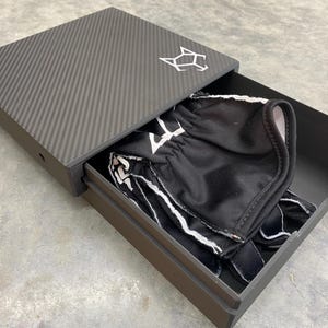 May include: A black carbon fiber box with a drawer that is open. The drawer contains a black and white patterned fabric item.