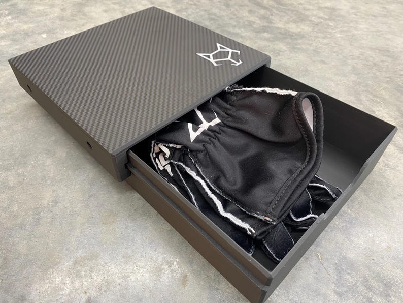 May include: A black carbon fiber box with a drawer that is open. The drawer contains a black and white patterned fabric item.