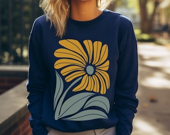 May include: A woman wearing a navy blue sweatshirt with a large yellow and green floral design. The flower has a yellow center and is surrounded by green leaves.