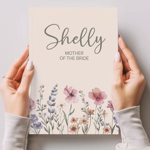 May include: A cream-coloured card held by hands, with the name "Shelly" in elegant script above "MOTHER OF THE BRIDE." The bottom edge features watercolour floral illustrations in shades of pink, purple, and yellow.