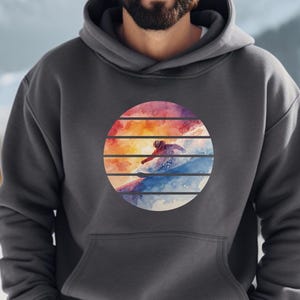 Könnte beinhalten: Ein anthrazitfarbener Kapuzenpullover mit einem kreisförmigen Grafikdesign eines Skifahrers auf einer schneebedeckten Piste, vor einem Sonnenuntergang in Orange, Rot und Blau. Der Hoodie hat eine Kapuze mit Kordelzug und eine Vordertasche.