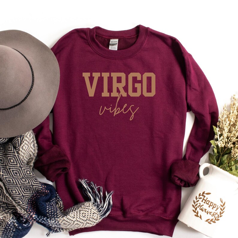 Pode incluir: Um moletom bord&ocirc; com gola redonda com as palavras "VIRGO vibes" impressas em dourado. O moletom &eacute; exibido com um chap&eacute;u cinza, um cachecol estampado e uma caneca branca com as palavras "Happy Harvest".