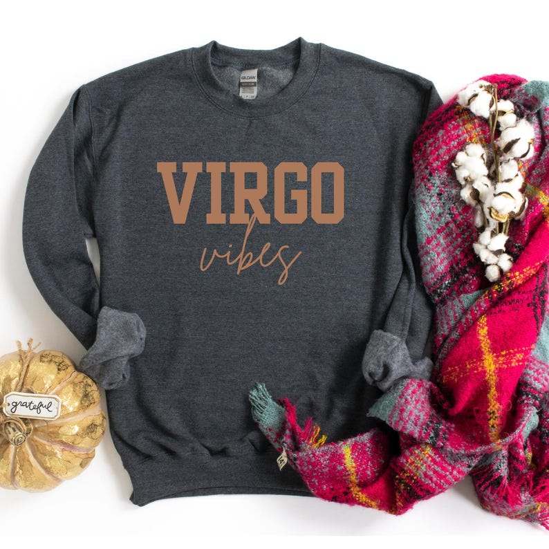 Pode incluir: Camisola cinza escuro com as palavras "VIRGO vibes" em letras castanhas. Uma ab&oacute;bora decorativa e um cachecol xadrez vermelho e rosa com caules de algod&atilde;o est&atilde;o ao lado da camisola.