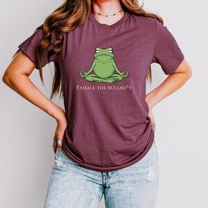 May include: A maroon t-shirt with a green frog meditating with the text "Exhale the Bullsh*t" printed on the front.