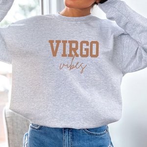 Pode incluir: Camisola cinza claro com a frase "VIRGO vibes" em castanho. A palavra "VIRGO" est&aacute; em letras de bloco, e "vibes" em it&aacute;lico.