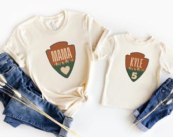 May include: Two cream colored t-shirts with a brown, green, and white park ranger badge design. The larger shirt says "MAMA" and the smaller shirt says "KYLE 5".