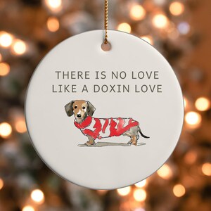 May include: A circular white ceramic Christmas ornament with a gold hanging string. It displays the text "THERE IS NO LOVE LIKE A DOXIN LOVE" and a watercolour illustration of a dachshund wearing a red and white scarf.