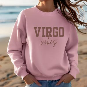 Pode incluir: Camisola rosa claro com o texto "VIRGO vibes" em letras castanhas. A camisola tem gola redonda e mangas compridas. A imagem &eacute; tirada numa praia.
