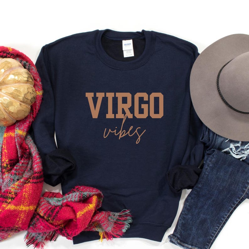 Pode incluir: Camisola azul marinho com as palavras "VIRGO vibes" impressas em castanho. Um chap&eacute;u cinzento, cal&ccedil;as de ganga azuis e um cachecol xadrez vermelho e amarelo s&atilde;o vis&iacute;veis. Parte de uma cole&ccedil;&atilde;o com tema de zod&iacute;aco.