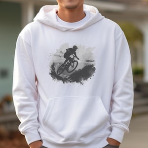 May include: White hoodie with a black and grey graphic of a mountain biker. The design depicts a cyclist riding a mountain bike downhill, set against a cloudy sky. The hoodie includes a front pocket and a drawstring hood.
