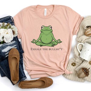 May include: A light pink t-shirt with a green frog meditating and the text "Exhale the Bullsh*t".