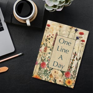Op de afbeelding: Een notitieboek met een bloemenpatroon en de tekst "One Line A Day" op de omslag. Het notitieboek ligt op een zwart oppervlak met een kopje koffie en een laptop.