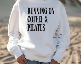 May include: White sweatshirt with the text "RUNNING ON COFFEE & PILATES" in black. The sweatshirt is long-sleeved and has a crew neck. The person is wearing blue jeans and has their hands in their pockets.