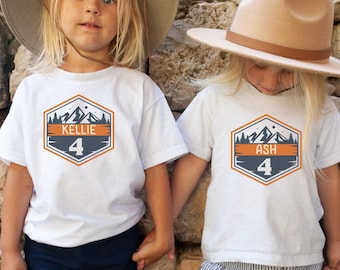May include: Two white t-shirts with a mountain and tree design. The shirts have the names "Kellie" and "Ash" with the number "4" on them.