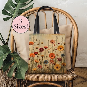 Könnte beinhalten: Tragetasche mit floralem Design mit orangefarbenen, roten, gelben und blauen Blumen. Die Tasche hat schwarze Griffe und den Text "3 Sizes!" in einem rosa Kreis. Die Tasche liegt auf einem Korbstuhl.