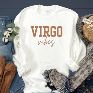 Pode incluir: Um moletom branco com gola redonda com as palavras "VIRGO vibes" impressas em marrom. O moletom &eacute; exibido com um par de jeans, um cachecol e uma ab&oacute;bora decorativa.