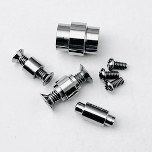 May include: An assortment of shiny, silver-colored metal hardware components. The image includes several screws, cylindrical pieces, and components with flared ends, all arranged against a white background. These parts appear to be precision-machined.