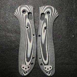 May include: Two knife handle scales with a black and white wood grain pattern. The scales have multiple holes for screws and pins. The design is symmetrical, with a curved shape for a comfortable grip. The scales are made of a durable material.