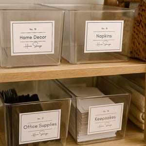Könnte beinhalten: Vier transparente Kunststoff-Aufbewahrungsboxen mit Etiketten mit der Aufschrift "Home Decor", "Napkins", "Office Supplies" und "Keepsakes". Die Boxen stehen auf einem hellen Holzregal. Die Etiketten haben den Text "Home Slange".