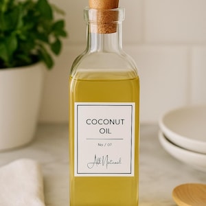 Pode incluir: Uma garrafa de vidro transparente cheia de óleo de coco amarelo, com uma rolha de cortiça e um bico dourado. O rótulo diz "COCONUT OIL" e "All Natural". A garrafa está sobre uma superfície de mármore.