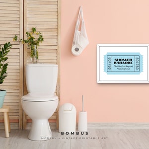 May include: A framed print with a light blue ticket design that reads "SHOWER KARAOKE" with the text "Birthday Suit Required" and "Talent optional". The print hangs on a pink wall above a toilet.