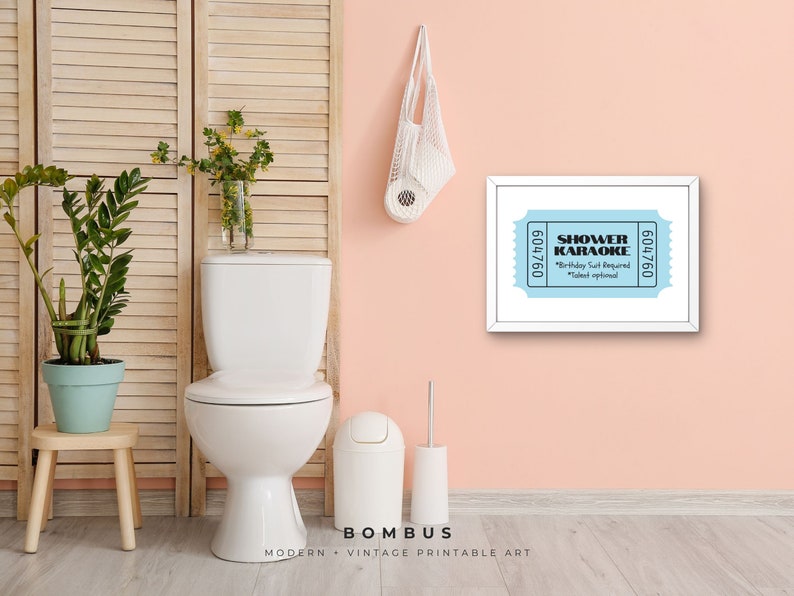 May include: A framed print with a light blue ticket design that reads "SHOWER KARAOKE" with the text "Birthday Suit Required" and "Talent optional". The print hangs on a pink wall above a toilet.