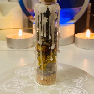 Puede incluir: Una pequeña botella de vidrio llena de hierbas secas, flores y cristales, coronada con un corcho y cera blanca. La botella está sobre una superficie blanca estampada, con velas encendidas en el fondo.