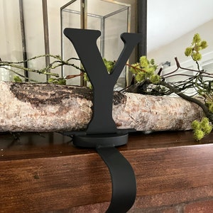 May include: A black metal stocking holder in the shape of the letter "Y". The holder features a curved arm for hanging a stocking. The letter sits on a circular base. The holder is positioned on a wooden mantelpiece.