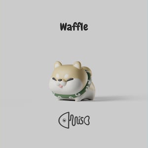 May include: A small, stylized figurine of a tan and white dog with a rounded body, wearing a green collar with a white swirl pattern. The word Waffle is printed in black above the figure, and a stylized fish logo is printed in black below.