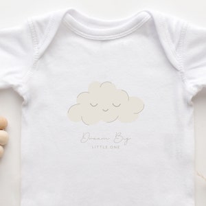 Puede incluir: Un body blanco para bebé con una nube marrón claro con una cara sonriente y el texto "Dream Big Little One" en gris.