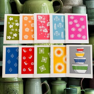 May include: A collection of colorful greeting cards with various retro floral and geometric designs. The cards feature white borders and vibrant backgrounds in shades of green, orange, pink, blue, and yellow. The cards are displayed in front of green teapots and ceramic containers.