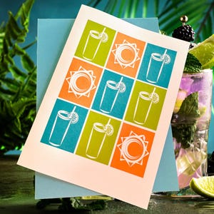 May include: A greeting card with a summer theme. The card features a grid of colorful squares with illustrations of cocktails and suns. The card is on top of a light blue envelope, with a cocktail in the background.