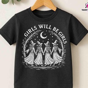 May include: Black t-shirt with the text "GIRLS WILL BE GIRLS" in a circular design. The graphic shows four figures in witch hats and dresses holding hands under a crescent moon and stars. The t-shirt is displayed on a wooden hanger.