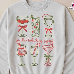May include: A light gray sweatshirt featuring a festive design with six cocktail glasses in red and green. The phrase "in the holiday spirit" is printed below the glasses. The design includes a bow, candy cane, and gingerbread man.