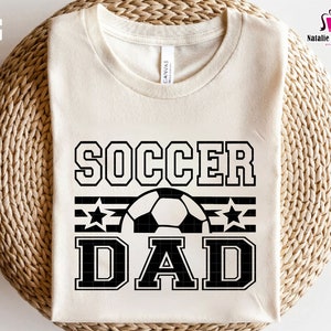 K&ouml;nnte beinhalten: Ein wei&szlig;es T-Shirt mit schwarzem Text, der "Soccer Dad" mit einem Fu&szlig;ball und Sternen lautet.