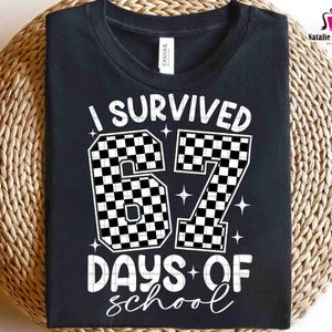 May include: Black t-shirt with the text "I Survived 67 Days of School" in white. The numbers "67" are designed with a black and white checkered pattern. The shirt is displayed on a woven surface.
