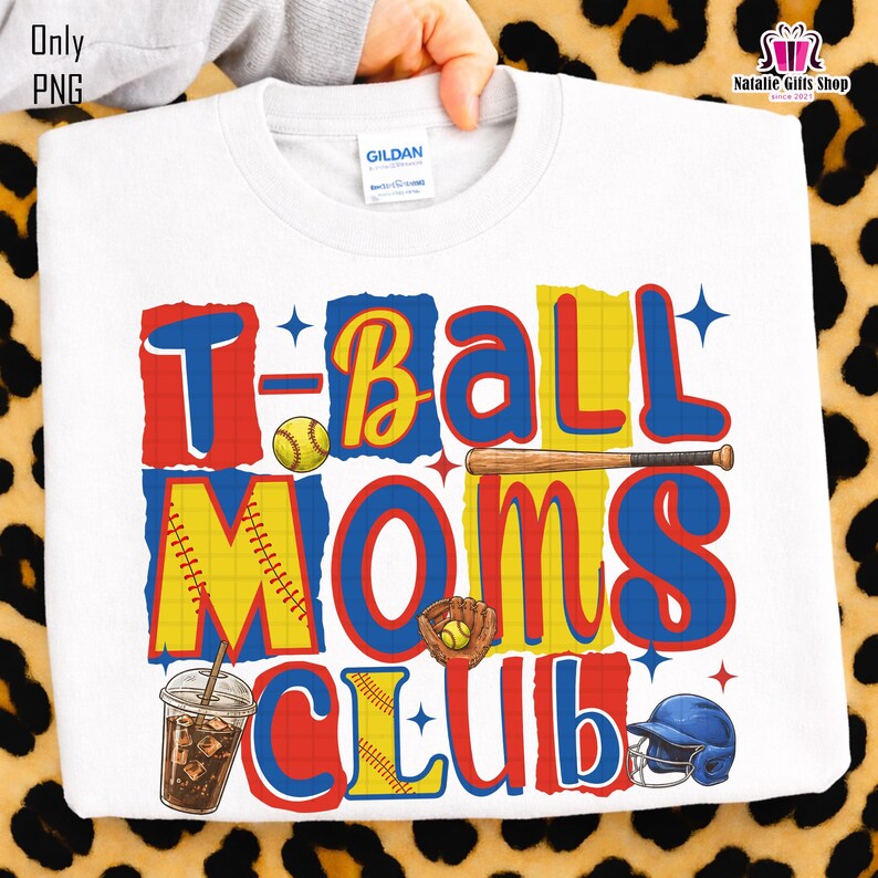 May include: White sweatshirt with the text "T-Ball Moms Club" in red, yellow, and blue. The design includes baseball-themed graphics such as a baseball, bat, glove, helmet, and a drink. The sweatshirt is held by a hand.