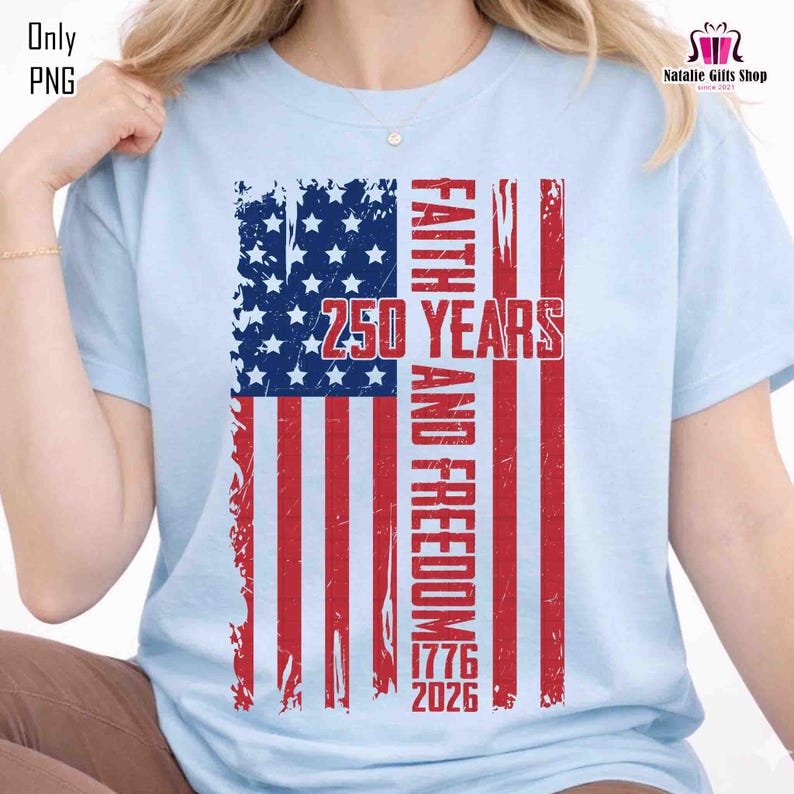 May include: Light blue t-shirt featuring a distressed American flag design. The flag incorporates the text "FAITH AND FREEDOM" and "250 YEARS" with the years "1776" and "2026". The shirt has short sleeves.