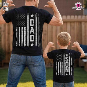May include: Black t-shirts with an American flag design. One shirt says "DAD" and the other says "SON" in white block letters. Both shirts have a distressed, vintage look. The shirts are worn by a father and son.
