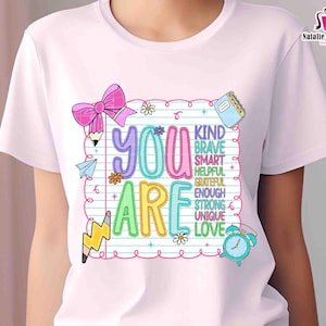 Puede incluir: Camiseta blanca con un dise&ntilde;o gr&aacute;fico colorido. El dise&ntilde;o incluye las palabras "YOU ARE" en letras grandes y contorneadas, con palabras adicionales como "KIND", "BRAVE" y "LOVE" dentro de un marco de papel rayado. Tambi&eacute;n se incluye un lazo rosa, l&aacute;pices y un reloj.