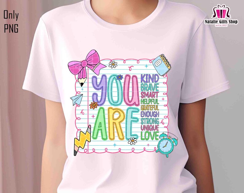Puede incluir: Camiseta blanca con un dise&ntilde;o gr&aacute;fico colorido. El dise&ntilde;o incluye las palabras "YOU ARE" en letras grandes y contorneadas, con palabras adicionales como "KIND", "BRAVE" y "LOVE" dentro de un marco de papel rayado. Tambi&eacute;n se incluye un lazo rosa, l&aacute;pices y un reloj.
